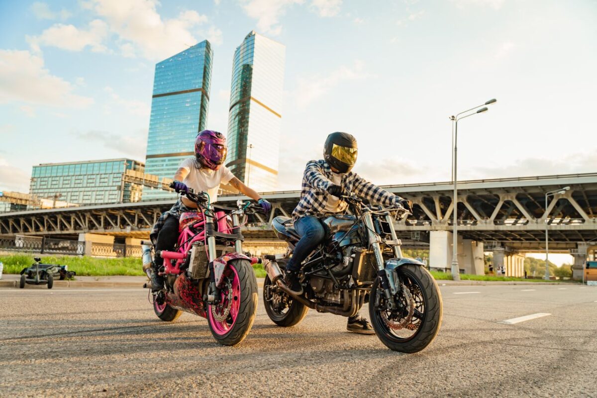 Simva Auto Two bikers riding motorcycles near urban skyscrapers at sunset.